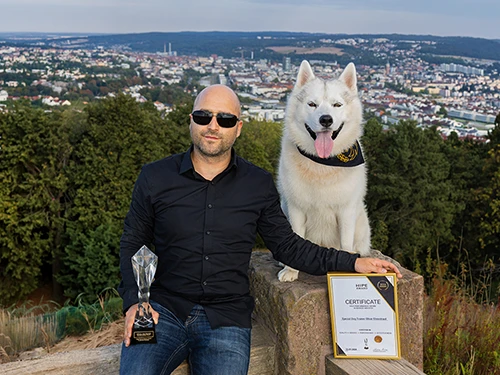 Special Dog Trainer Oliver Ehrentraut und sein weißer Husky Kuma posieren draußen glücklich mit dem HIPE-Award und der zugehörigen Auszeichnungsurkunde.